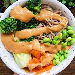 Hand holding white bowl with soba noodles, veggies and edamame with peanut lime sauce on top