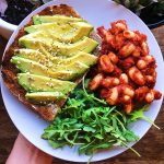 Hand holding a blue plate with avocado toast, baked beans and arugula salad.
