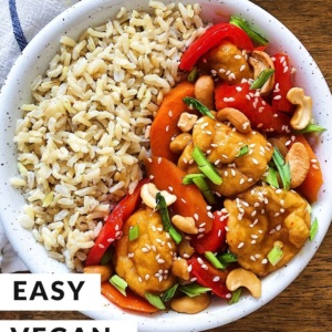 Cashew tofu with bell peppers and sliced green onion along with brown rice in a bowl on a wooden board with a napkin.
