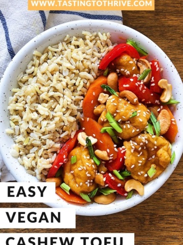 Cashew tofu with bell peppers and sliced green onion along with brown rice in a bowl on a wooden board with a napkin.