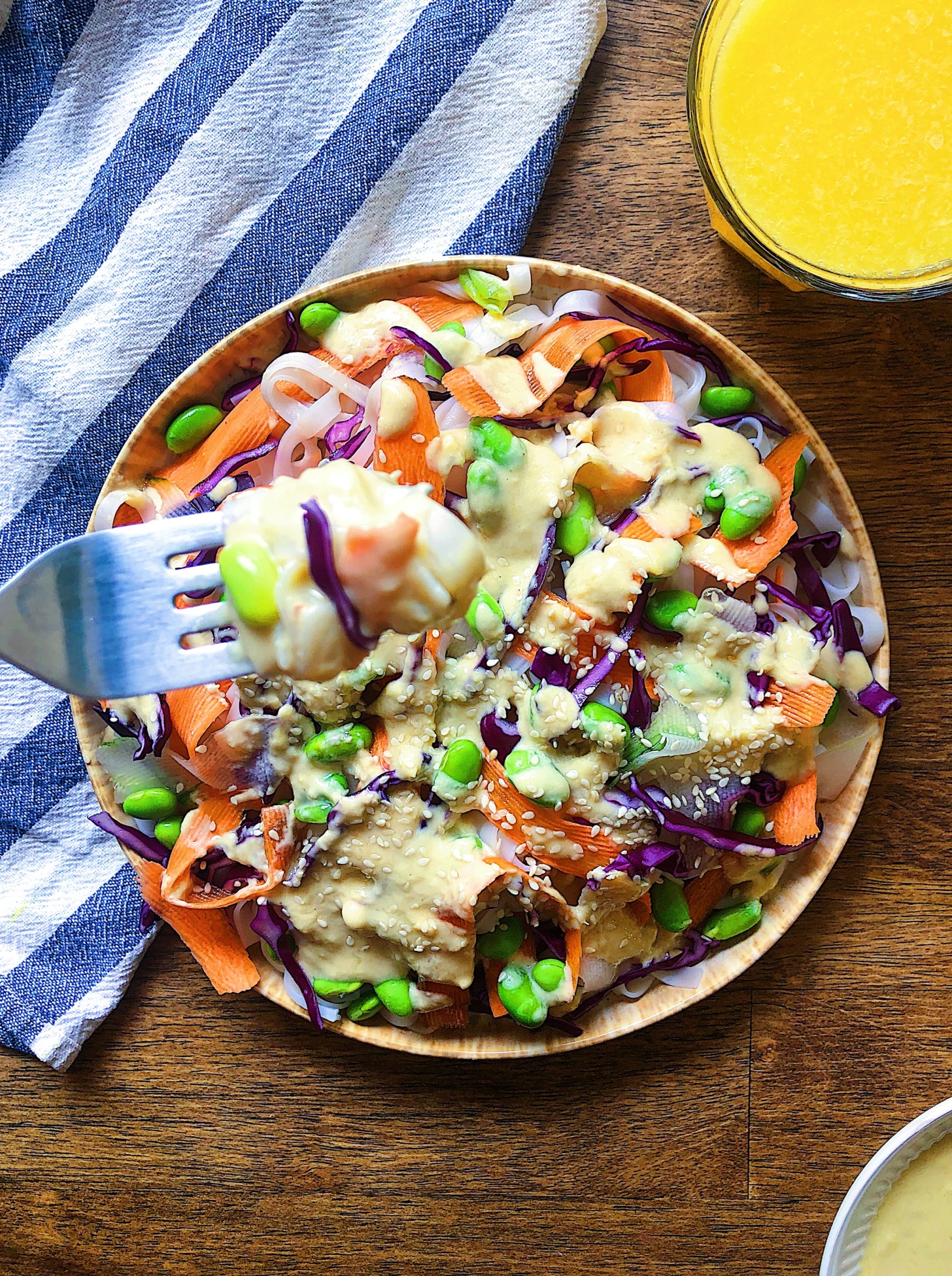 Rice noodles on a plate with edamame, sliced carrots, purple cabbage and a sauce over top with a fork scooping some and orange juice to the side.