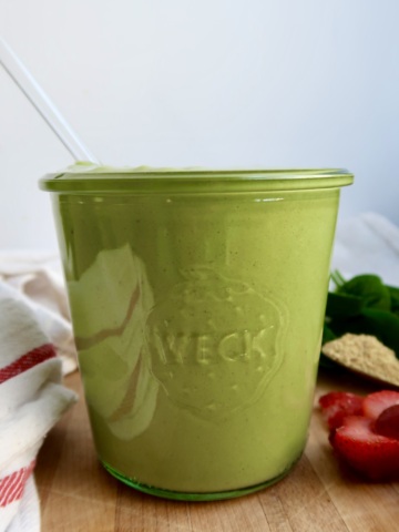 Jar with green smoothie with strawberries and spinach in the background.