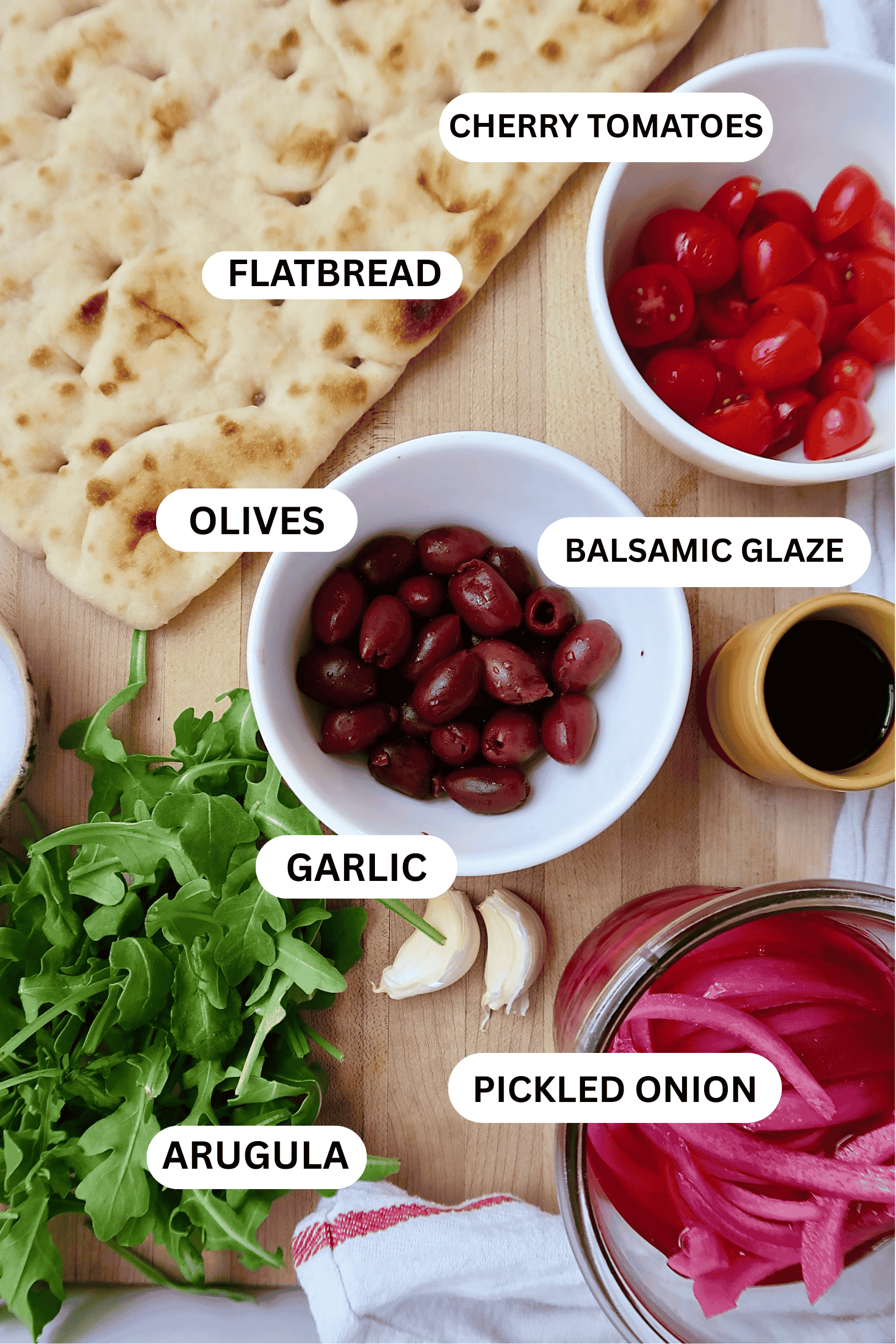 Ingredient flatlay with flatbread, cherry tomato, kalamata olive, pickled red onion, arugula, garlic, pickled onion.