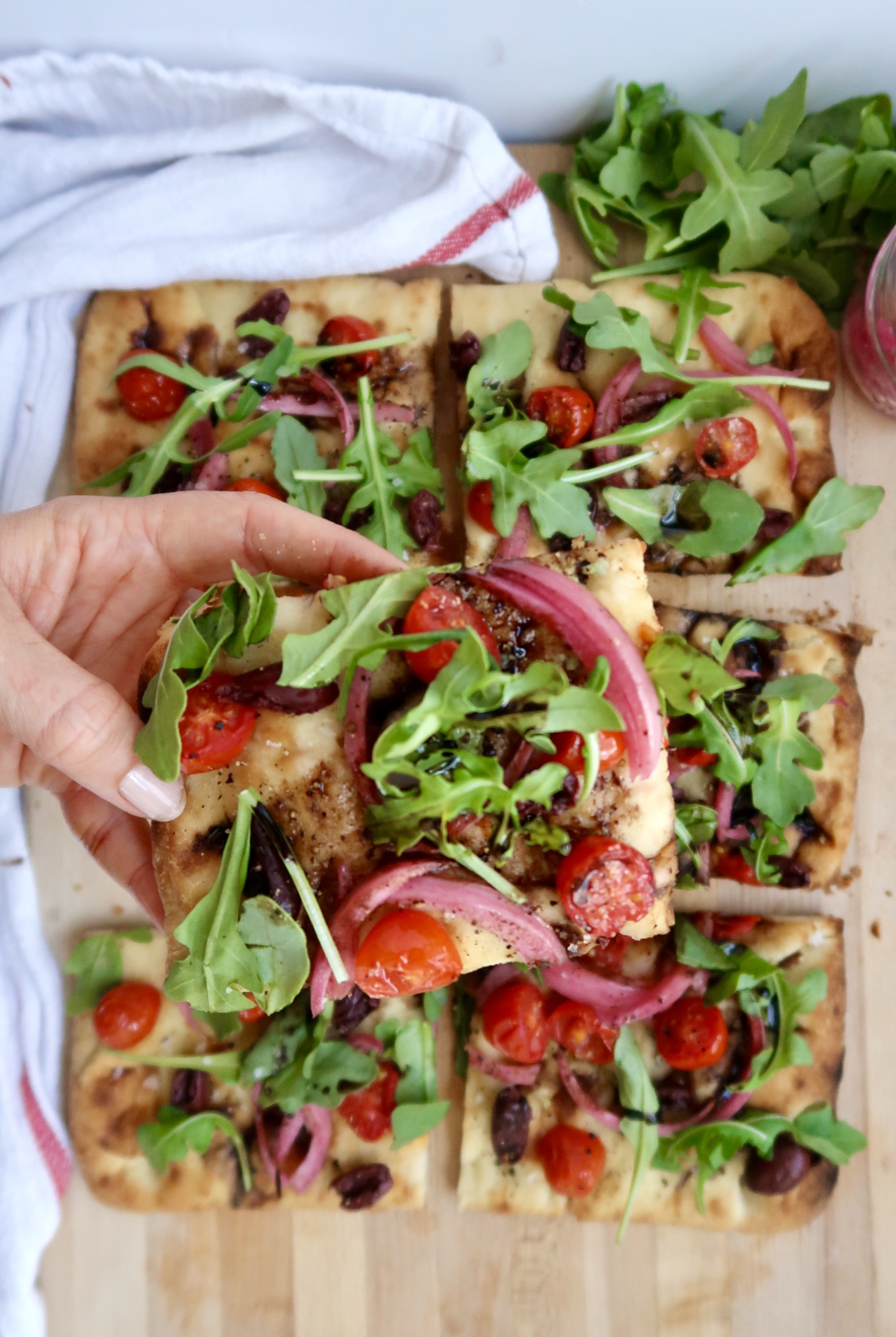 Flatbread with cherry tomato, olives and arugula and balsamic glaze, sliced, and hand holding a piece.