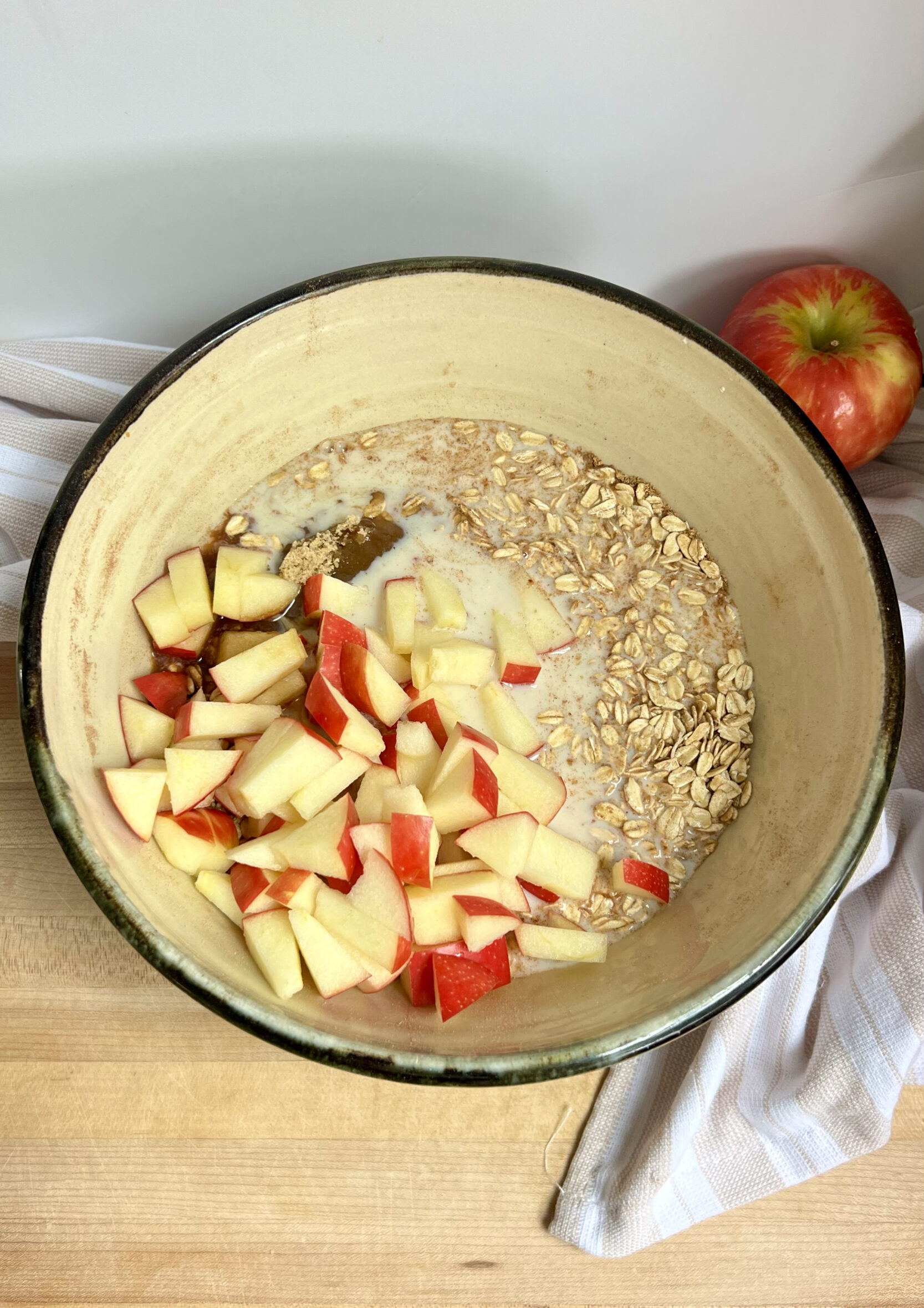 Large bowl with oats, soy milk, sliced apple inside on top of a cutting board.