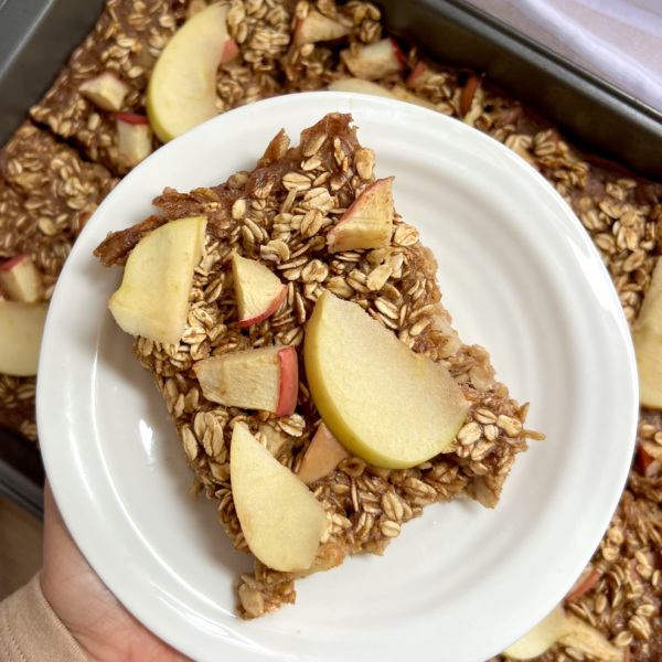 Hand holding a white plate of baked oats with sliced apples on top
