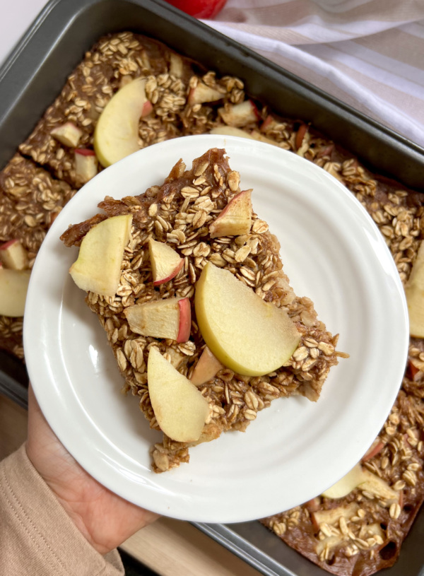 Hand holding a white plate of baked oats with sliced apples on top