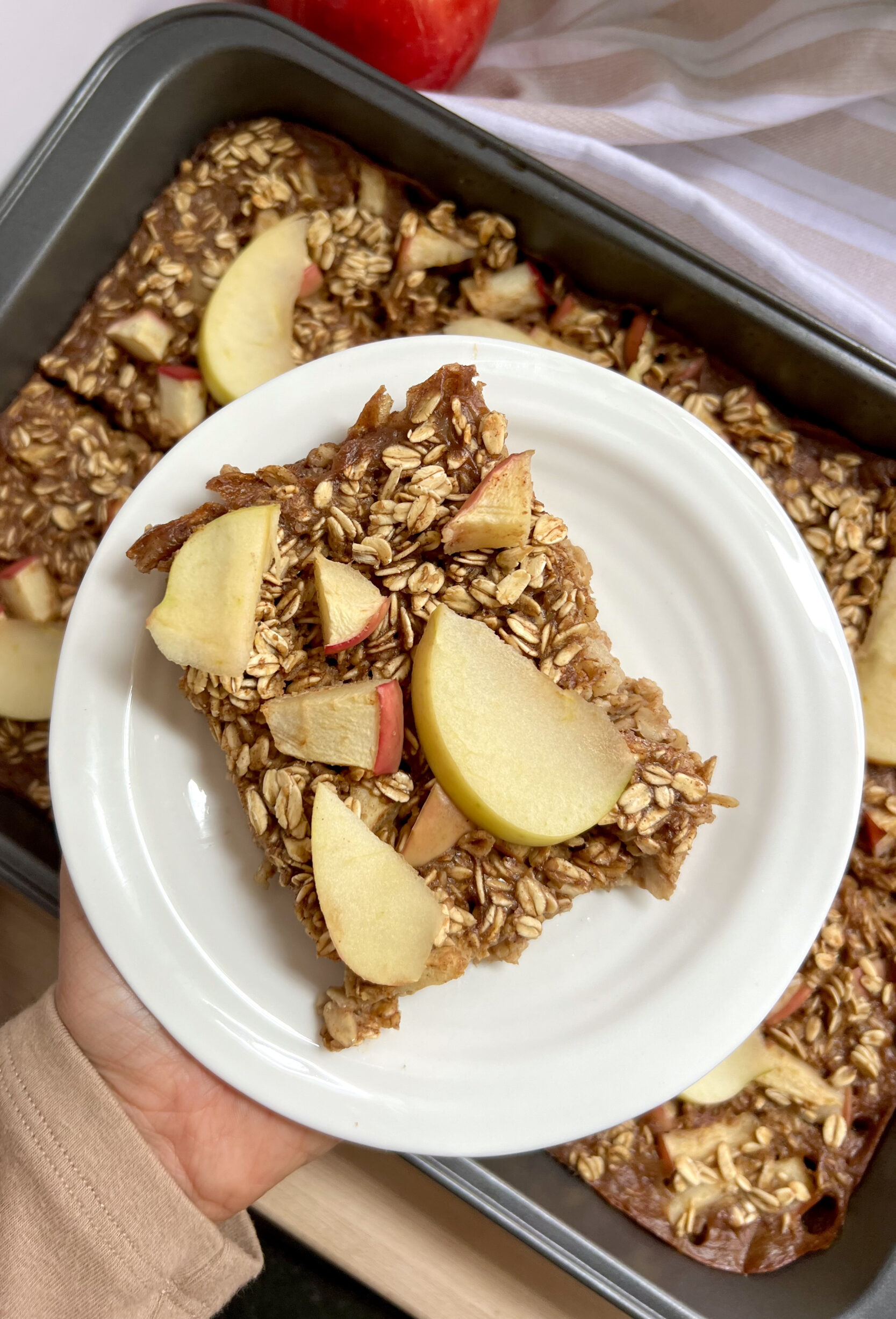 Hand holding a white plate of baked oats with sliced apples on top
