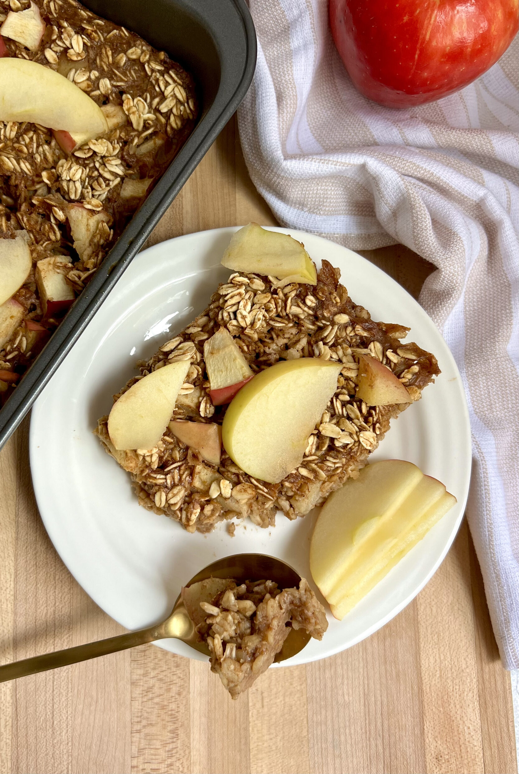 Cutting board with a white plate of baked oats on top with a tray of it to the side and sliced apple on top.