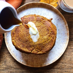 Plate with a stack of pancakes on it with a slab of butter on top and pouring maple syrup over top.