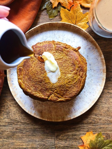 Plate with a stack of pancakes on it with a slab of butter on top and pouring maple syrup over top.