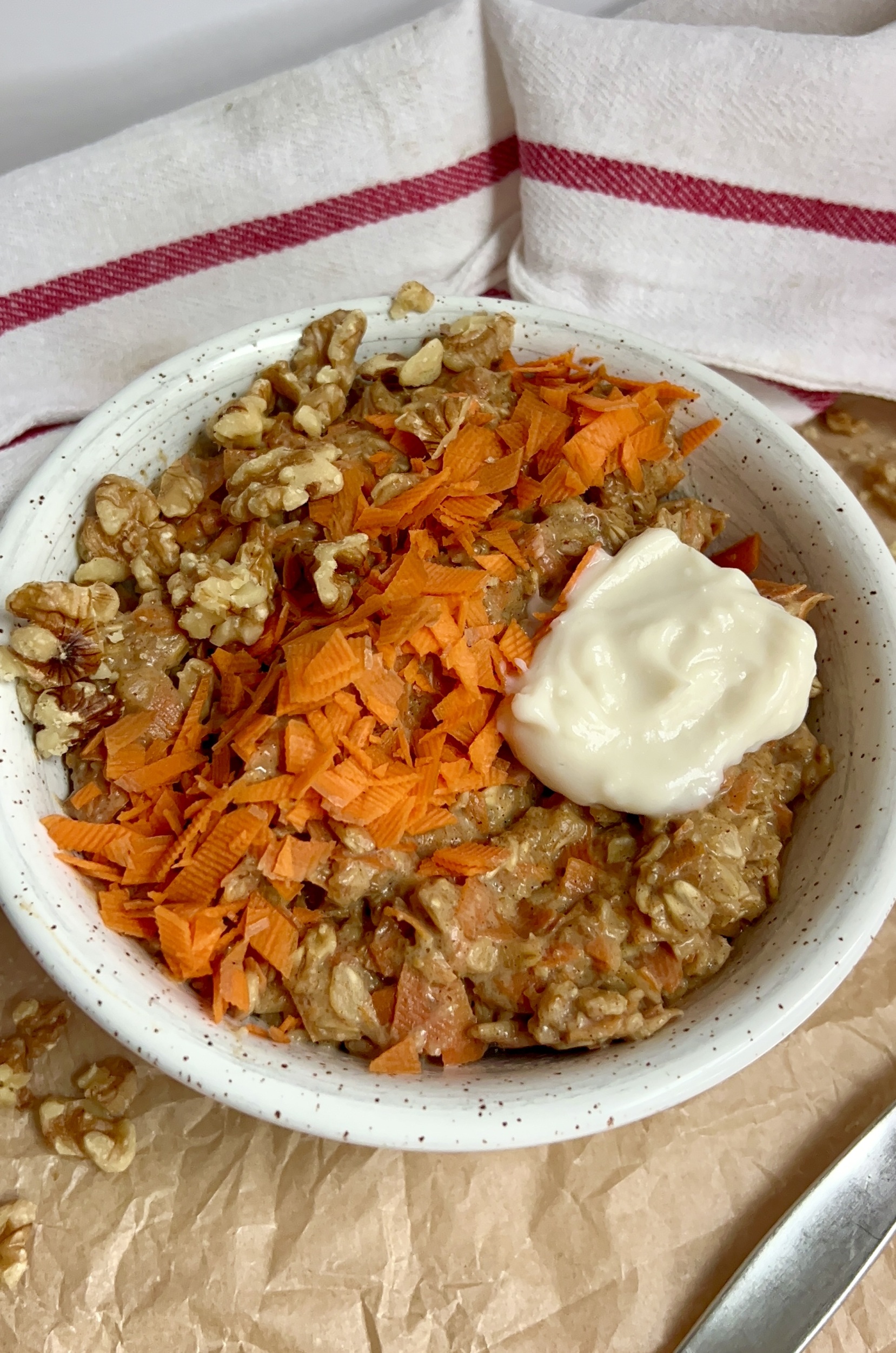 White bowl on parchment paper with oatmeal, grated carrot, walnuts on top with a spoon to the side and a dollop og yogurt on top.