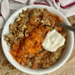 White bowl on parchment paper with oatmeal, grated carrot, walnuts on top with a dollop of yogurt on top and a spoon in the oatmeal.