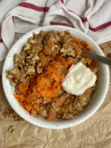 White bowl on parchment paper with oatmeal, grated carrot, walnuts on top with a dollop of yogurt on top and a spoon in the oatmeal.
