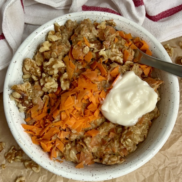 White bowl on parchment paper with oatmeal, grated carrot, walnuts on top with a dollop of yogurt on top and a spoon in the oatmeal.
