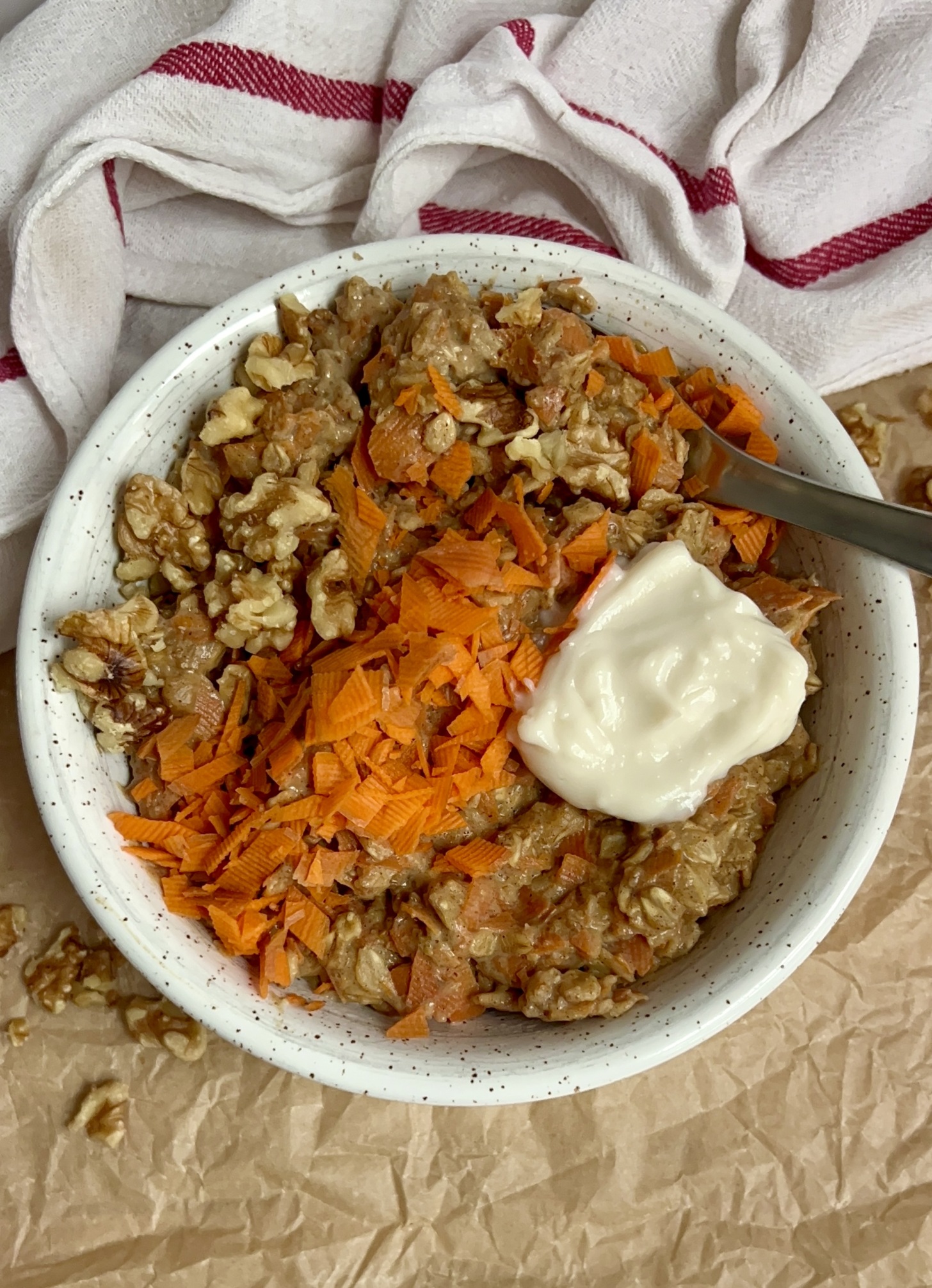 White bowl on parchment paper with oatmeal, grated carrot, walnuts on top with a dollop of yogurt on top and a spoon in the oatmeal.