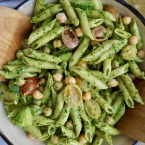 A bowl of penne pasta with green pesto sauce, cherry tomatoes, chickpeas, and chopped vegetables, being served with wooden utensils.