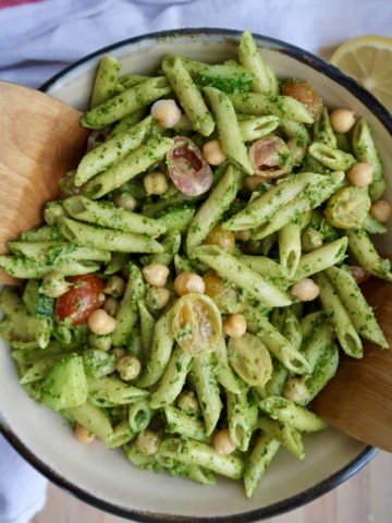 A bowl of penne pasta with green pesto sauce, cherry tomatoes, chickpeas, and chopped vegetables, being served with wooden utensils.
