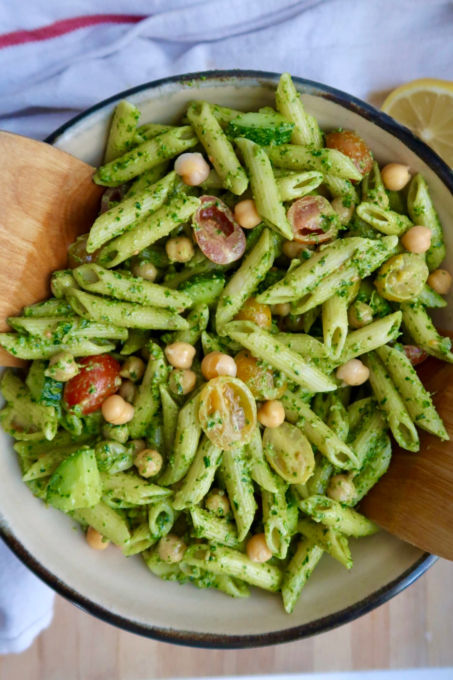 A bowl of penne pasta with green pesto sauce, cherry tomatoes, chickpeas, and chopped vegetables, being served with wooden utensils.