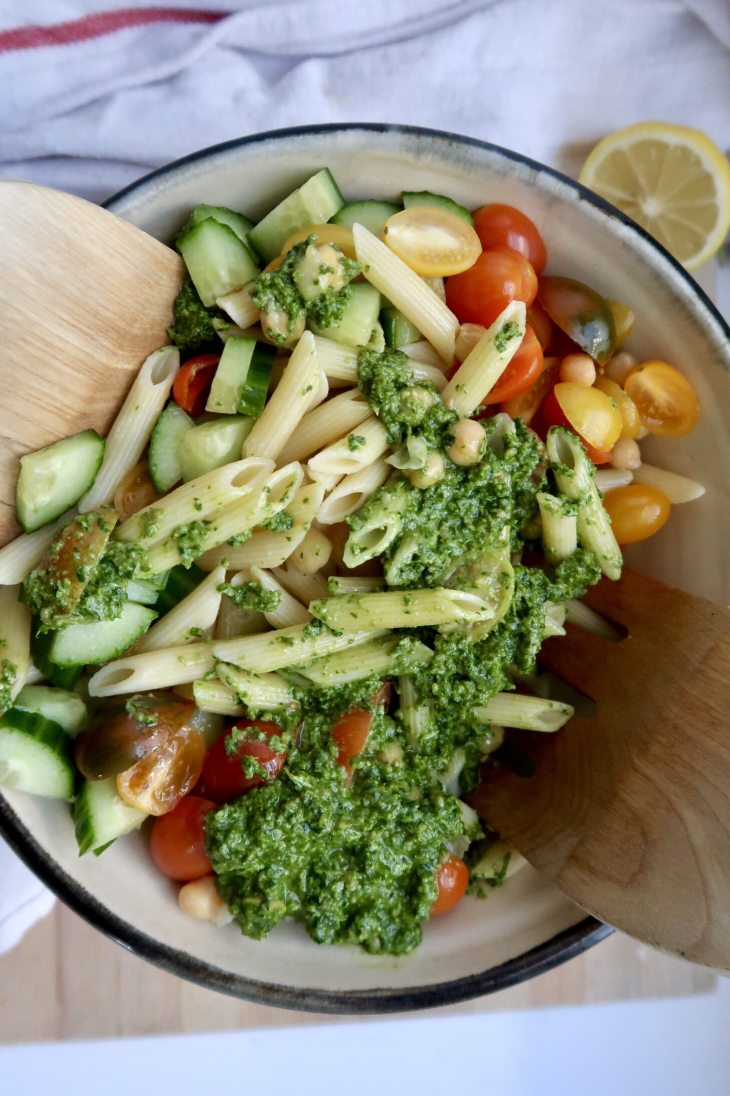 A bowl of penne pasta salad with cherry tomatoes, cucumber, chickpeas, and a generous dollop of green pesto, being mixed with two wooden utensils. A lemon slice and a towel are partially visible in the background.