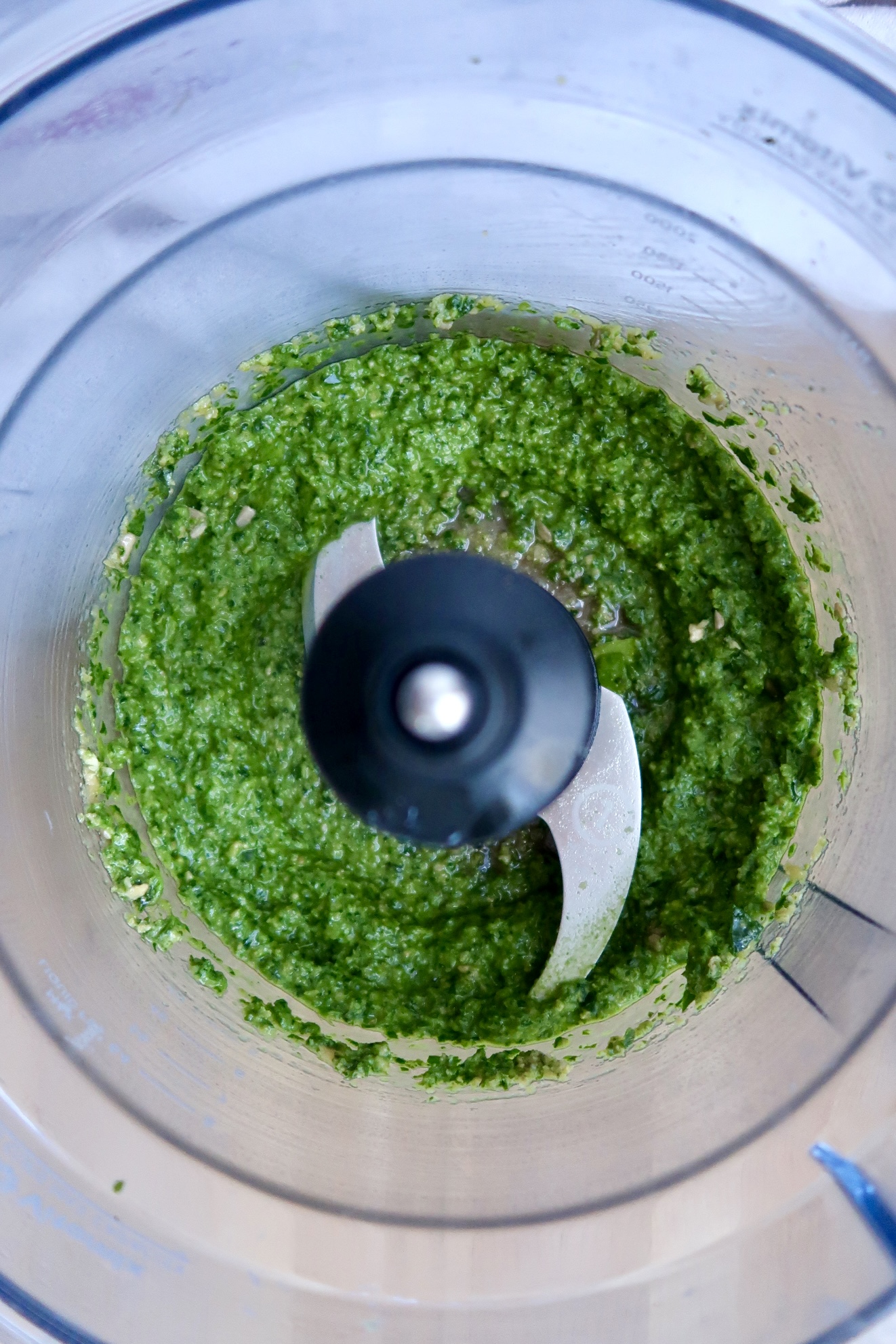 A top-down view of a food processor containing freshly blended green pesto, with the metal blades visible in the center.