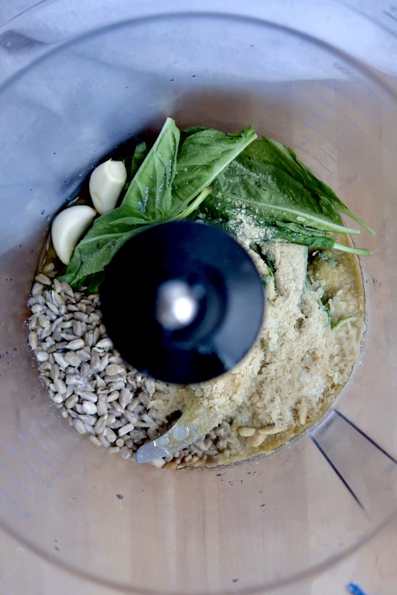 Overhead view of a food processor containing fresh basil leaves, garlic cloves, sunflower seeds, nutritional yeast, and oil, ready to be blended into a pesto sauce.