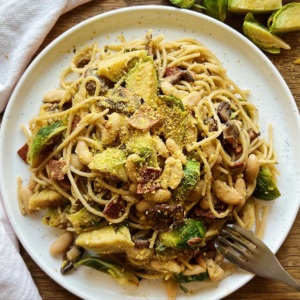 White plate with spaghetti with roasted brussel sprouts, bacon and white beans with nutritional yeast on top.