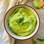 Bowl of basil edamame hummus with basil lead on top on a white background and a cracker dipped in the hummus in the corner