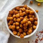 White background with small white bowl of Chili Lime Roasted Chickpeas with lime on the side to garnish.