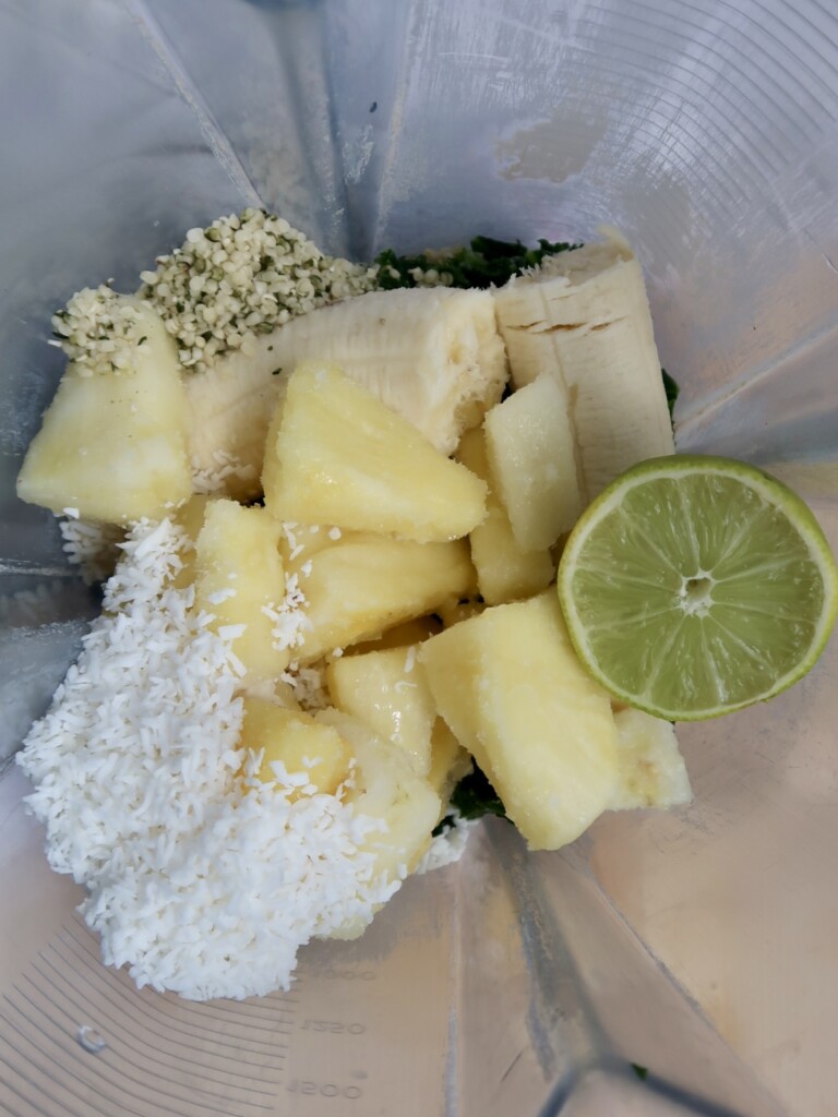 Kale, pineapple, coconut flakes, banana, lime, hemp seeds and soy milk in a smoothie jug.