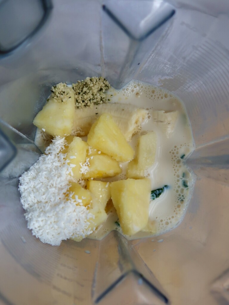 Kale, pineapple, coconut flakes, hemp seeds and soy milk in a smoothie jug.
