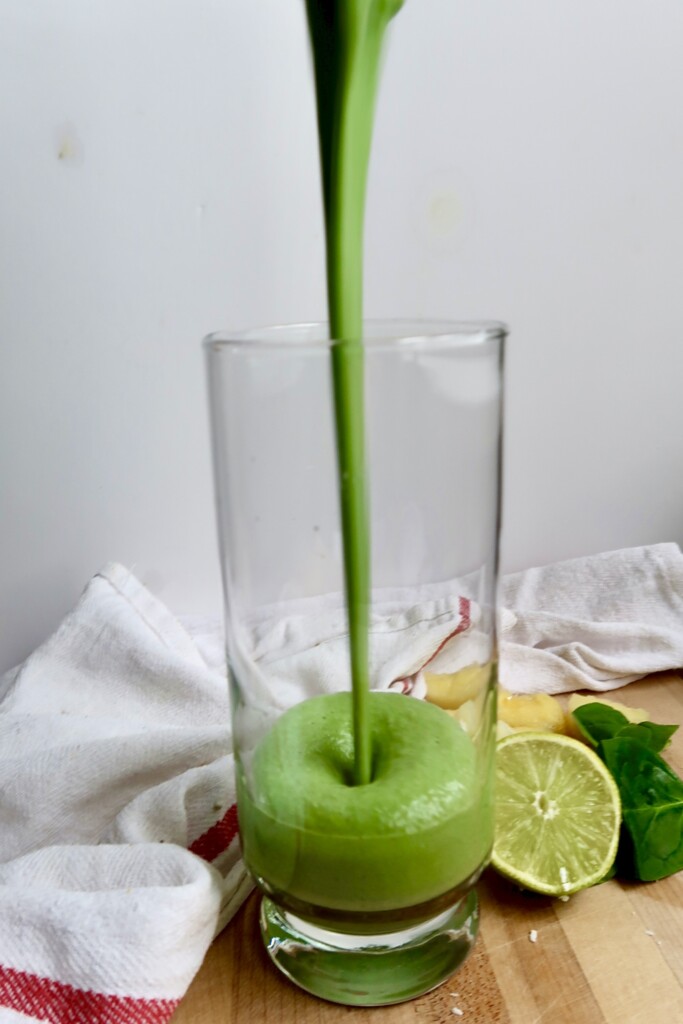 Glass with a green smoothie being poured into it and fresh lime on the side.