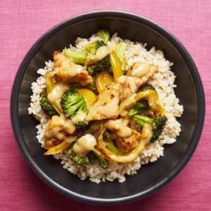 Pink background with a black bowl with rice, broccoli, bell pepper and miso dijon tempeh with a sauce drizzled on top.