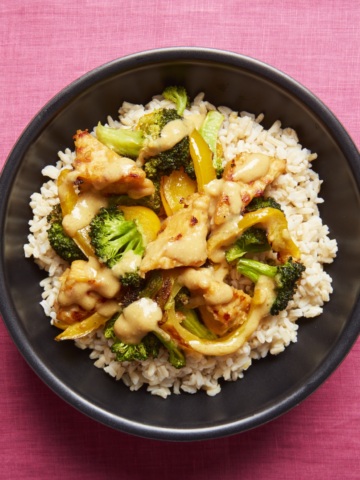 Pink background with a black bowl with rice, broccoli, bell pepper and miso dijon tempeh with a sauce drizzled on top.