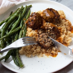 White plate with vegan meatballs, couscous and garlic green beans. Fork and knife cutting through the vegan meatball.