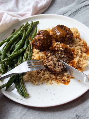 White plate with vegan meatballs, couscous and garlic green beans. Fork and knife cutting through the vegan meatball.
