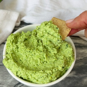 Small white bowl with cilantro lime edamame dip with a cracker dipping into it.