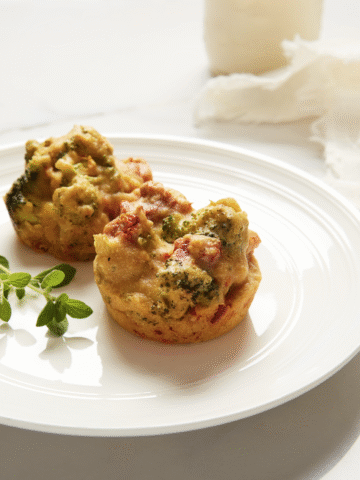 Two egg bites on a white plate that have sundried tomatoes and broccoli in them with greenery to the side.