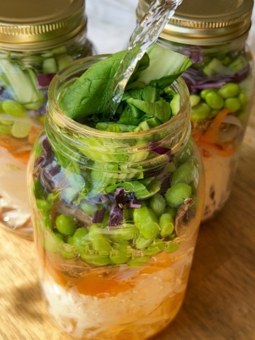 Three large mason jars layered with rice noodles, sliced carrot, edamame, bok choy, shredded purple cabbage and hot water being poured in.