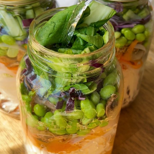 Three large mason jars layered with rice noodles, sliced carrot, edamame, bok choy, shredded purple cabbage and hot water being poured in.