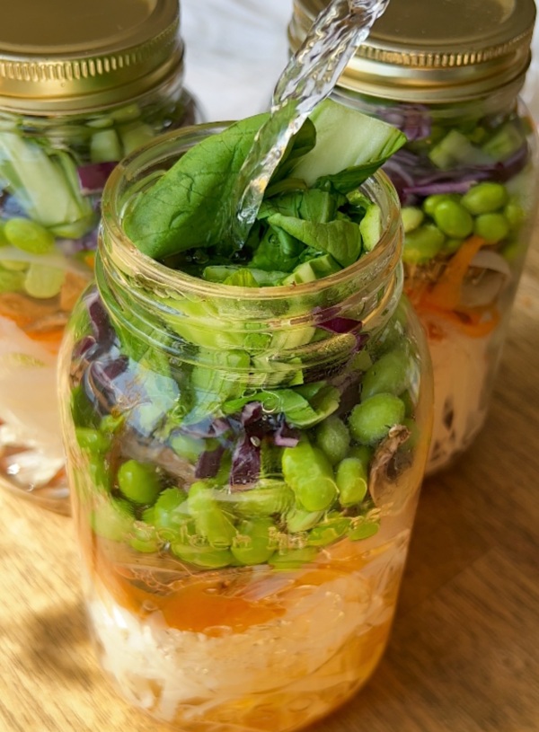 Three large mason jars layered with rice noodles, sliced carrot, edamame, bok choy, shredded purple cabbage and hot water being poured in.