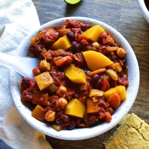 Bowl of butternut squash chili with chunks of butternut squash in a white bowl, cornbread and lime in the corner and a white and blue napkin under the bowl.