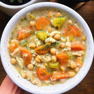 Hand holding a white bowl filled with soup with carrots, celery, pasta and chickpeas in it.