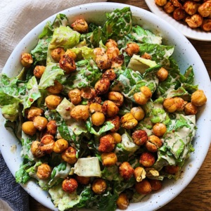 White bowl with caesar salad and crispy chickpeas on top, with a small bowl of crispy chickpeas on the side and a white and blue napkin next to it.