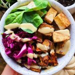 White bowl with garlic tofu, purple cabbage sweet potato, spinach and a creamy sauce on top, with plants in the background