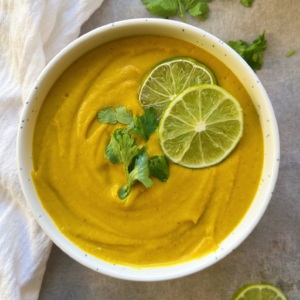 White background with a white bowl with orange coloured sweet potato, carrot and ginger soup that's blended with lime and cilantro to garnish.