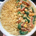 Hand holding white bowl with quinoa in one half and a sundried tomato and coconut milk braised chickpea curry with spinach. Plants in the background.