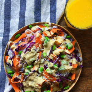 Plate with noodles, shaved carrot, edamame, and cabbage with an orange miso dressing drizzled on top. Glass of orange juice next to the plate and a white and blue striped napkin.