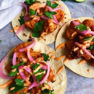 White background with three tacos with cauliflower, pickled onions, sriracha mayo and cilantro on top.