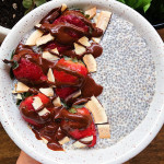 Hand holding white bowl with chia pudding in it as well as strawberries, coconut flakes, and drizzled with chocolate.